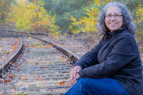 Smiling mature woman with grayish black hair and glasses, sitting on train tracks, autumnal trees in the Meinweg nature reserve, old Iron Rhine railway (IJzeren Rijn) in Middle Limburg, Netherlands photo