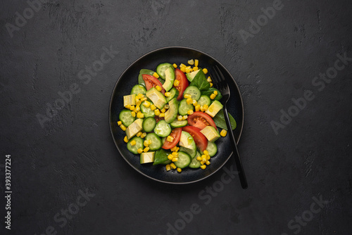Avocado salad with corn, tomatoes, cucumbers on black slate background. Top view, copy space