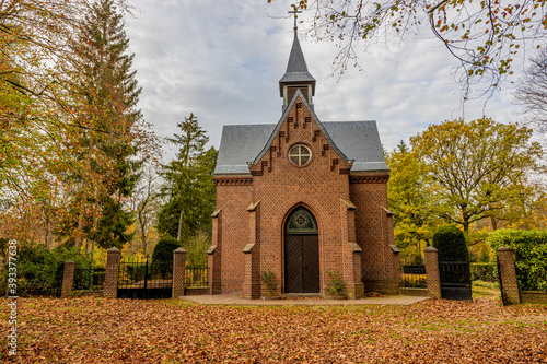 St. Ludwig Chapel surrounded by autumnal trees with yellowish green leaves, the ground covered with fallen brown leaves, cloudy day in the Meinweg nature reserve in Middle Limburg, the Netherlands