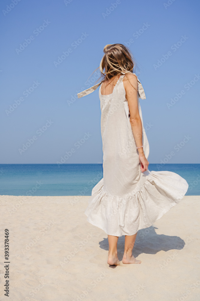 A blonde smiling girl in a maxi linen dress with fluttering hair jumping and dancing on a sand beach against blue sky and sea background