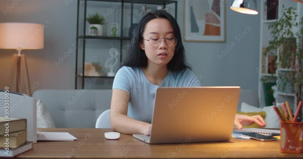 Portrait of Asian young woman in glasses texting on smartphone and typing on laptop while sitting in room indoor alone. Busy female at workplace working on computer. Employee concept Portrait of Asian young woman in glasses texting on smartphone and typing on laptop while sitting in room indoor alone. Busy female at workplace working on computer. Employee concept