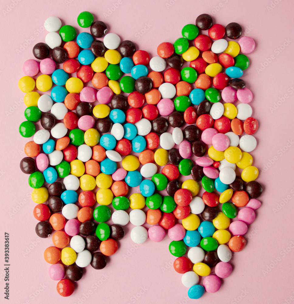 Obraz premium Tooth of chocolate multicolored candies flat lay view on pink background. Tooth made from candies.