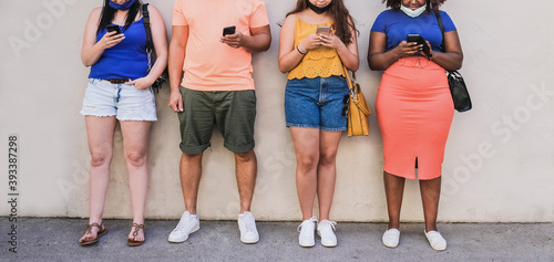 Young multiracial people standing in a line and look at their mobile phones while wearing protective face mask under chin - Coronavirus lifestyle © Sabrina