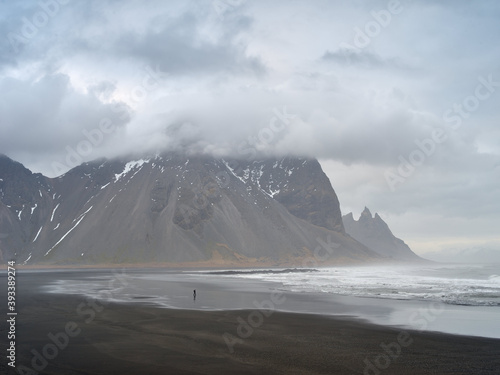 A tourst walking on the beach of Stokksnes, Mount Vestrahorn in background. Gray cloudy mood. Iceland.