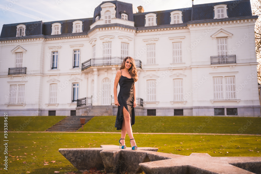 Lifestyle, blonde girl in a glamorous dress and blue high heels in a white house. Elegant outfit in autumn at sunset next to a palace in the background