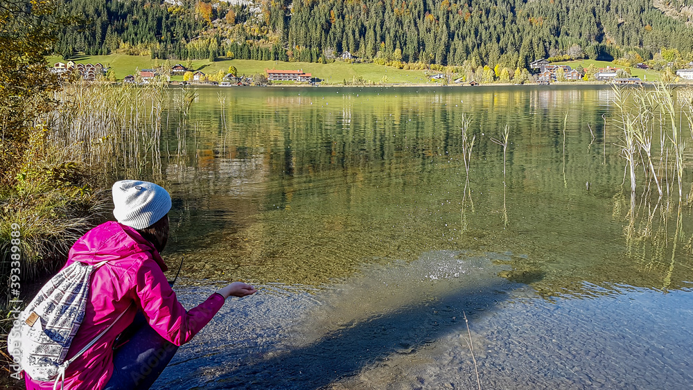Obraz premium A woman squatting at the side of Weissensee and playing with the cold water. She wears a pink jacket and a beanie. The surface of the lake is calm and reflecting the Alps. Joyfulness and happiness.
