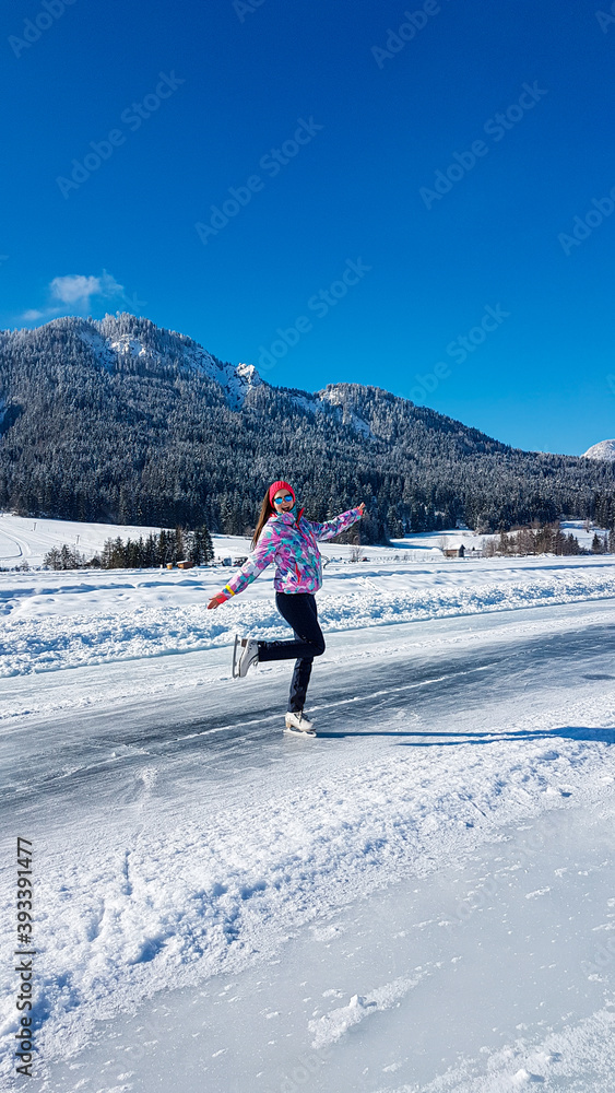 A woman in colorful outfit skating on a frozen Weissensee lake in Austria. The lake is surrounded by mountains. The ice rink is well prepared. Winter activity. Winter wonderland. Happiness