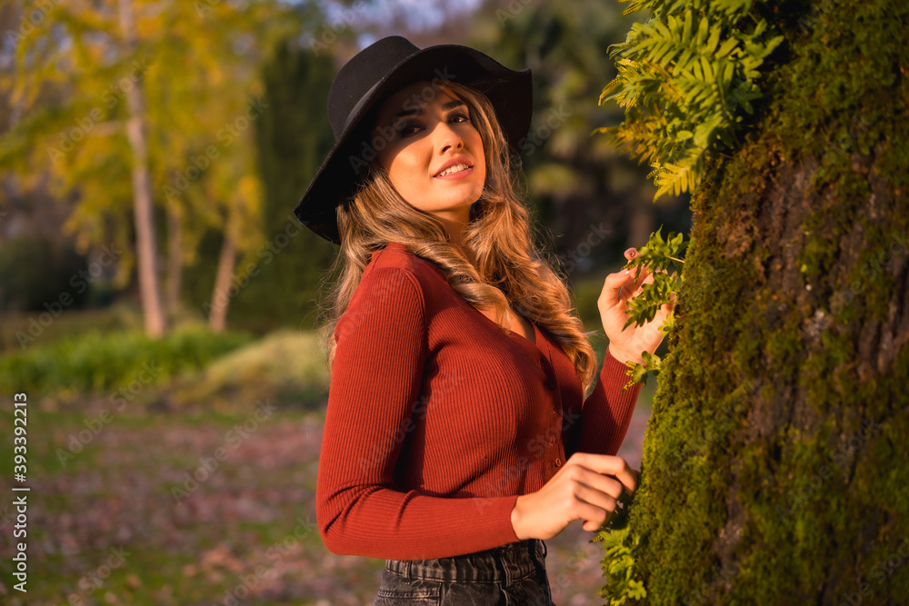 Lifestyle, blonde Caucasian girl in a red sweater and a black hat, enjoying nature in a park with trees, portrait of the attractive young woman in autumn