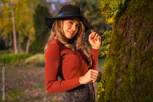 Lifestyle, blond Caucasian girl in a red sweater and a black hat, enjoying nature in a park with trees in the beautiful autumn, portrait of the young woman next to a tree with moss photo