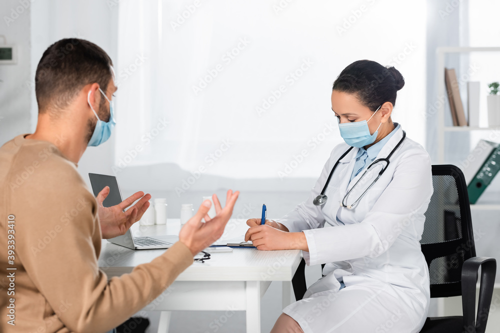 Fototapeta premium African american doctor writing on clipboard, while patient gesturing in clinic on blurred background