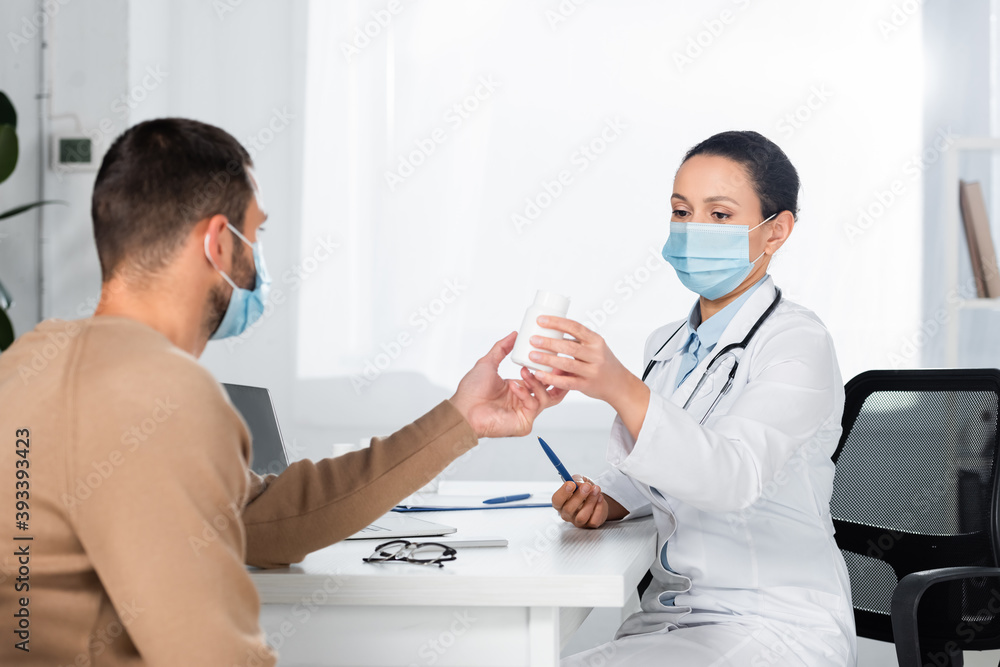 African american doctor giving bottle with pills to patient, while sitting at workplace on blurred background