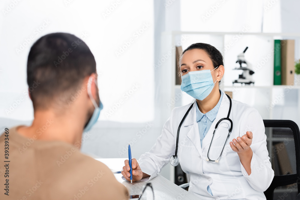 African american doctor in medical mask gesturing and looking at patient, while sitting at desk on blurred background African american doctor in medical mask gesturing and looking at patient, while sitting at desk on blurred background