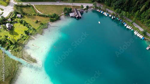 A top down, drone capture of Weissensee lake in Austrian Alps. The water changes color from white, turquoise to navy blue. A small settlement at the shore. A bit of overcast. Serenity and peacefulness