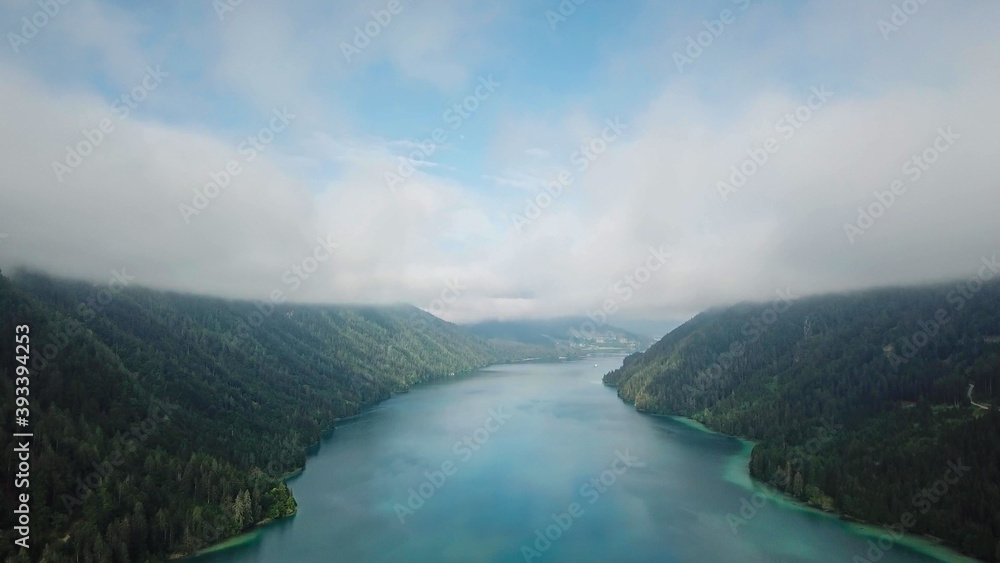 Idyllic, drone capture of Weissensee lake in Austria. The lake is surrounded by high Alps. The forest overgrown the slopes. The water has turquoise color. A bit of overcast. Serenity and peacefulness