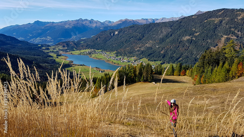 A woman in pink jacket enjoying the Alpine landscape while hiking with the view on Weissensee lake. The grass is golden, trees also change colors to yellow and orange. Autumn vibes photo