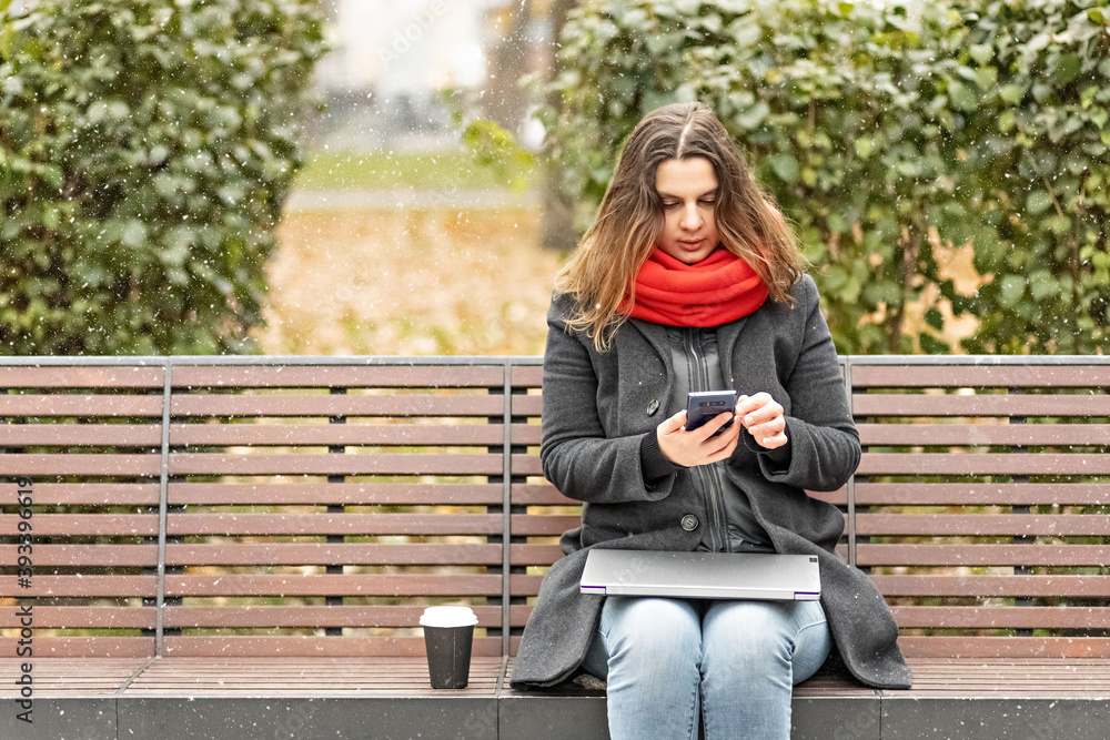 Young woman holds a smartphone and checks messages while sitting on a park bench with a laptop on her lap. Young woman holds a smartphone and checks messages while sitting on a park bench with a laptop on her lap.