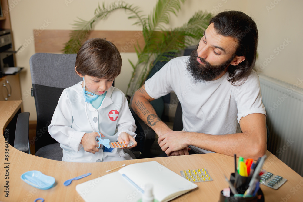 Little boy playing pretends like doctor examining a man in comfortabe medical office. Healthcare, childhood, medicine, protection and happiness concept. Having fun, laughting while giving pills Little boy playing pretends like doctor examining a man in comfortabe medical office. Healthcare, childhood, medicine, protection and happiness concept. Having fun, laughting while giving pills