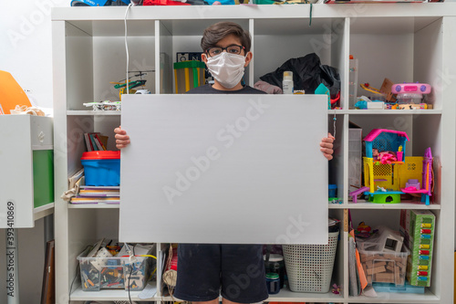 Covid-19 Stay at home concept: A little boy with glasses wears a face mask and holds a white empty board without a message. The kid stands in front of a toy shelf in the children room