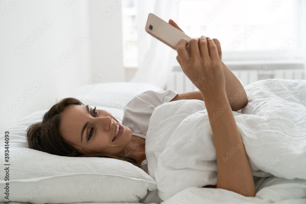 Female enjoying lazy weekend morning. Smiling millennial young woman using mobile smart phone, lying and relaxing in white comfortable bed at home, chatting in social networks. Female enjoying lazy weekend morning. Smiling millennial young woman using mobile smart phone, lying and relaxing in white comfortable bed at home, chatting in social networks.