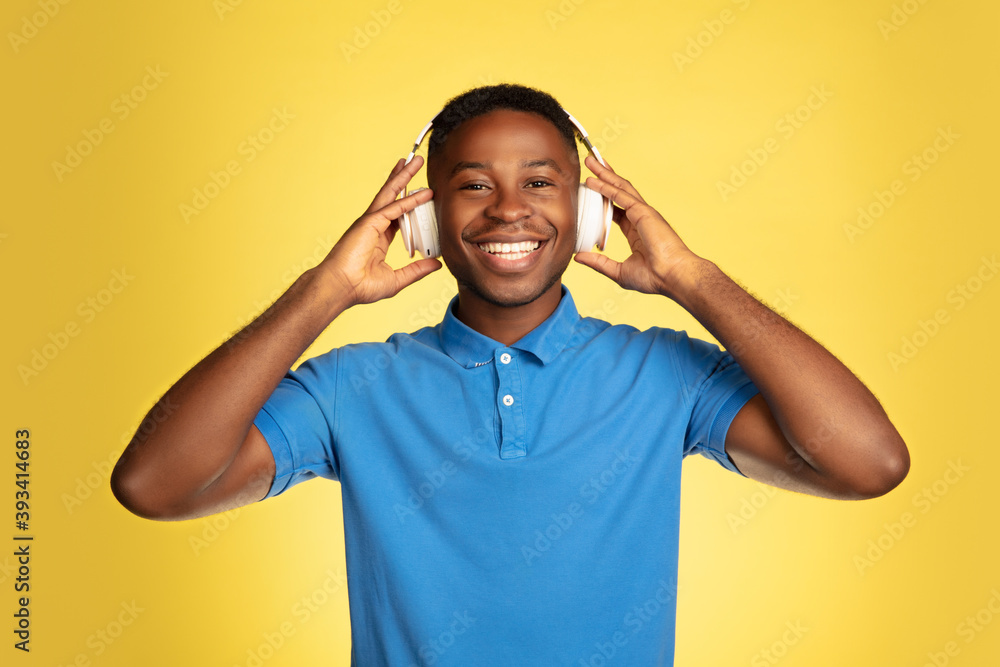 Music in headphones. Young african-american man's portrait isolated on yellow studio background, facial expression. Beautiful male portrait with copyspace. Concept of human emotions, facial expression Music in headphones. Young african-american man's portrait isolated on yellow studio background, facial expression. Beautiful male portrait with copyspace. Concept of human emotions, facial expression