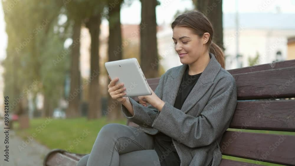 Slow Motion Young girl sits on the bench in autumn city square and looks and flips through the photos in his tablet computer Slow Motion Young girl sits on the bench in autumn city square and looks and flips through the photos in his tablet computer