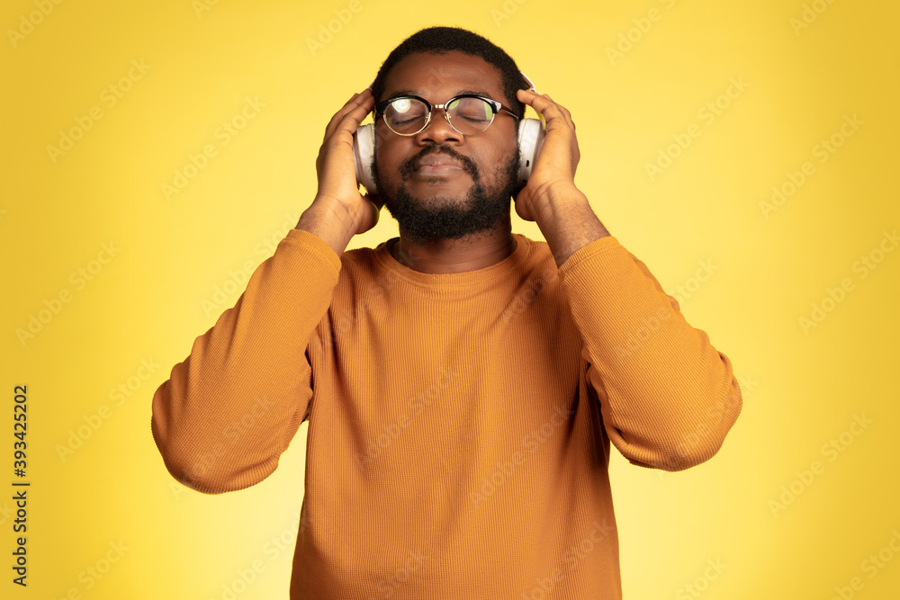 Enjoying. Young african-american man's portrait isolated on yellow studio background, facial expression. Beautiful male model with headphones, copyspace. Concept of human emotions, sales, ad. Enjoying. Young african-american man's portrait isolated on yellow studio background, facial expression. Beautiful male model with headphones, copyspace. Concept of human emotions, sales, ad.