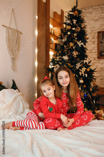 Two cute little sisters in red pajamas are sitting on the bed and hugging against the background of a Christmas tree with garlands and lights. Christmas and new year holidays concept with family