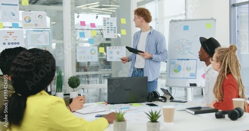 Attractive positive successful creative guy with curly hair standing near glass wall with diagramms and explaining to his mixed race colleagues strategy of further joint work