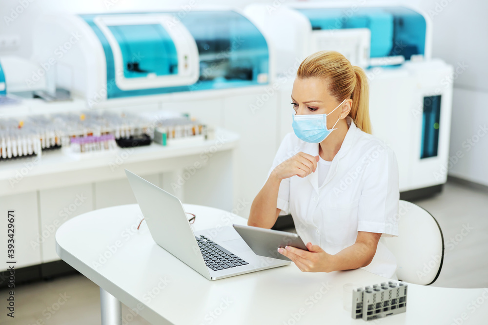 Experienced lab assistant sitting in laboratory with face mask on, holding tablet with test results and looking at laptop. Research for cure for corona virus concept. Experienced lab assistant sitting in laboratory with face mask on, holding tablet with test results and looking at laptop. Research for cure for corona virus concept.