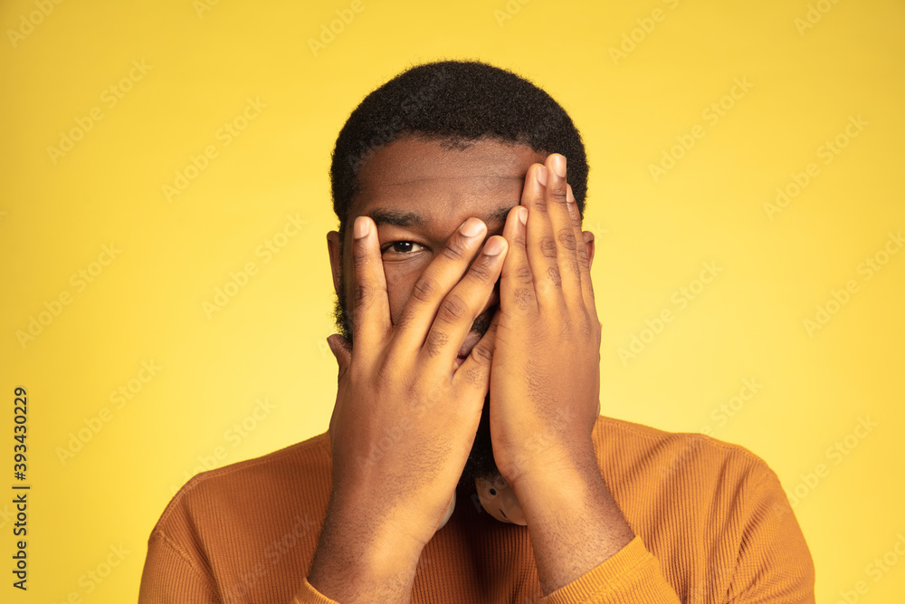 Scared hiding face. Young african-american man's portrait isolated on yellow studio background, facial expression. Beautiful male model with headphones, copyspace. Concept of human emotions, sales, ad Scared hiding face. Young african-american man's portrait isolated on yellow studio background, facial expression. Beautiful male model with headphones, copyspace. Concept of human emotions, sales, ad