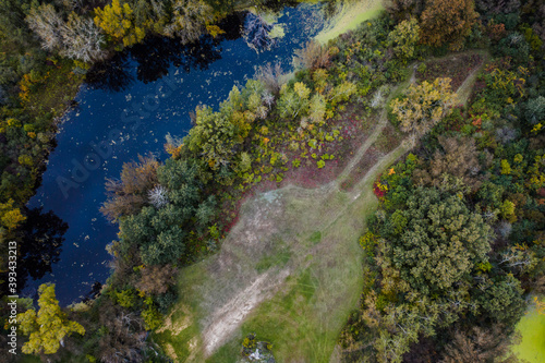 Aerial look down photograph of a pond surrounded by green and yellow trees with an open grass area with a worn trail through a portion of the wooded area in Wisconsin.