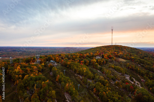 Beautiful aerial drone photograph of rib mountain with colorful fall leaves or autumn foliage lining the grassy ski runs at sunset as the cloudy sky turns orange and pink above the horizon.