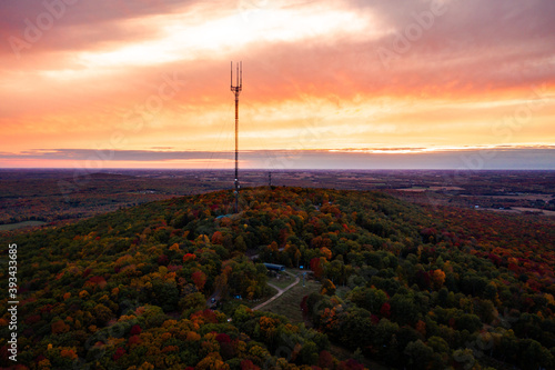Beautiful aerial drone photograph of rib mountain as the sky erupts with orange and pink colored clouds beyond the broadcast tower at sunset with colorful fall leaves or autumn foliage on hill below.