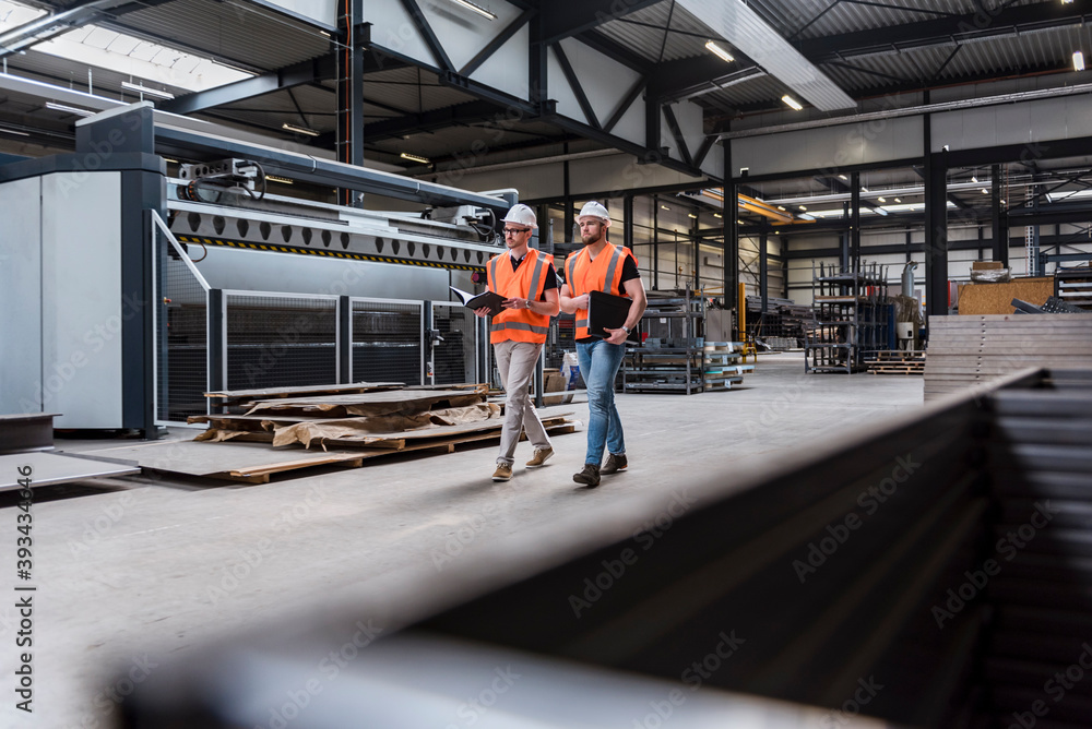 Two men wearing hard hats and safety vests walking on factory shop floor Two men wearing hard hats and safety vests walking on factory shop floor