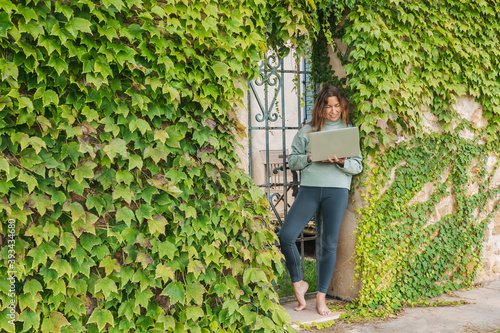 Mature woman using laptop while leaning on ivy wall at gate