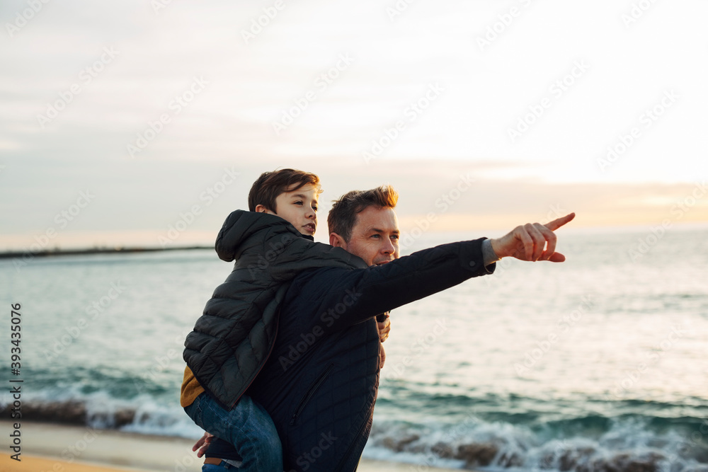 Father carrying son piggyback on the beach at sunset pointing finger Father carrying son piggyback on the beach at sunset pointing finger