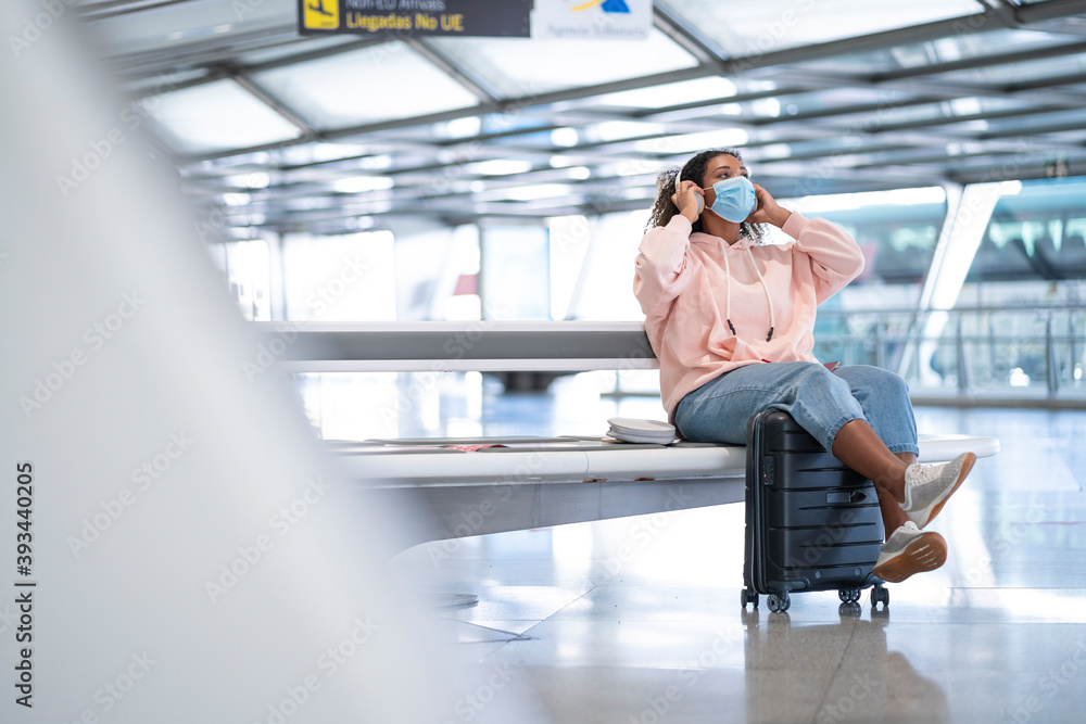 Young woman listening to music while wearing protective face mask sitting at airport