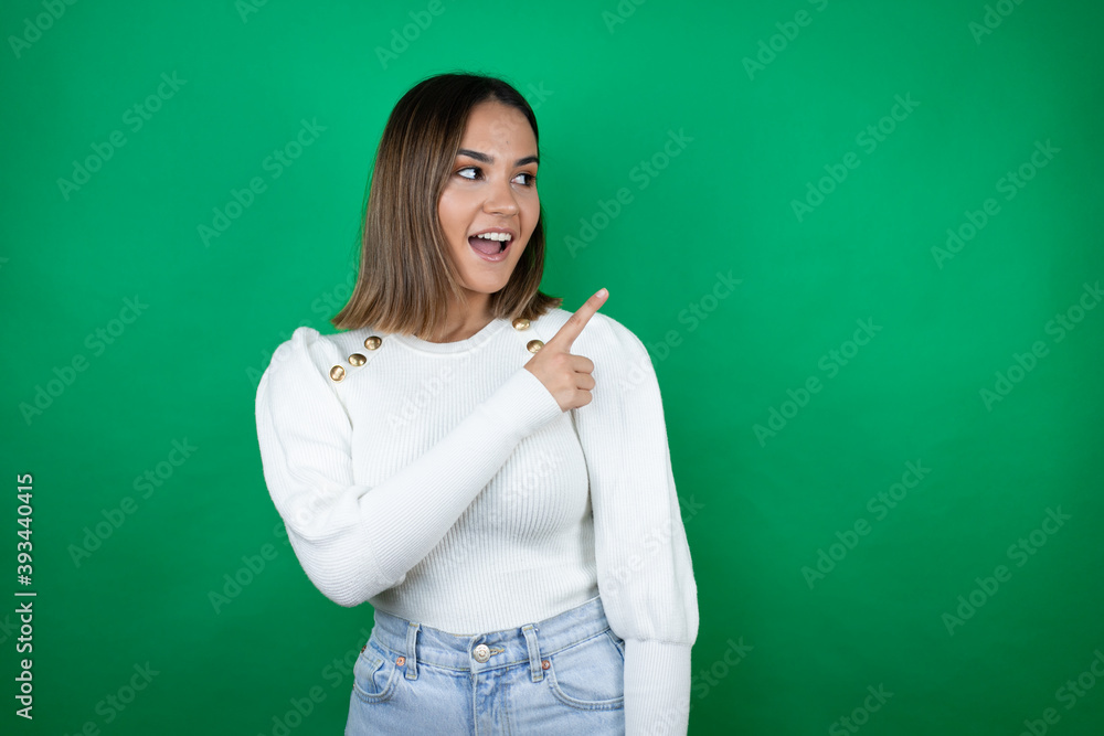 Young beautiful woman wearing white sweater over isolated green background surprised and pointing with hand and finger to the side Young beautiful woman wearing white sweater over isolated green background surprised and pointing with hand and finger to the side