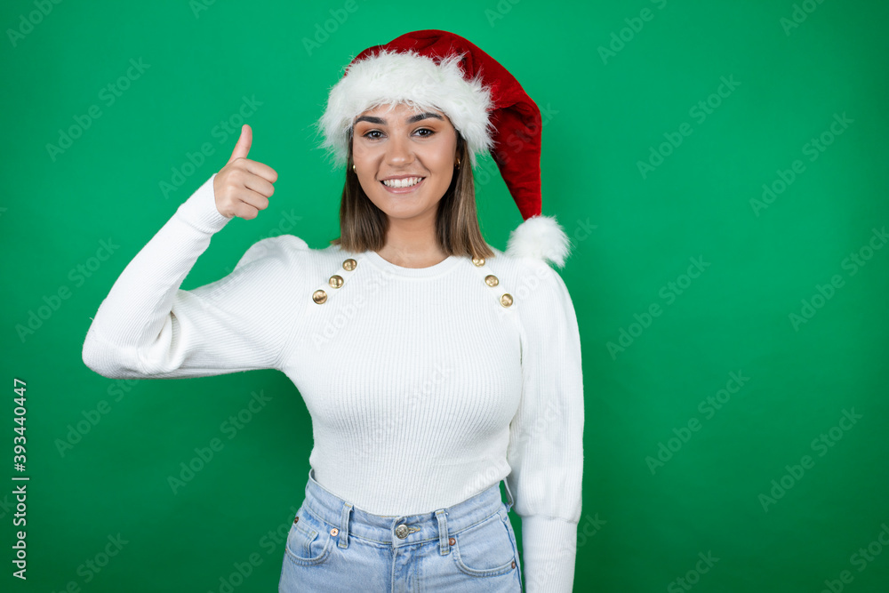 Young beautiful woman wearing a Santa hat and white sweater over green background smiling and doing the ok signal with her thumb Young beautiful woman wearing a Santa hat and white sweater over green background smiling and doing the ok signal with her thumb
