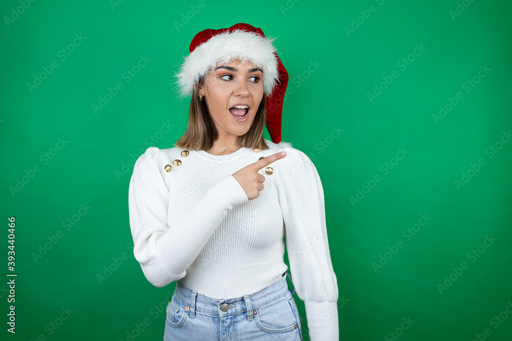 Young beautiful woman wearing a Santa hat and white sweater over green background surprised and pointing with hand and finger to the side