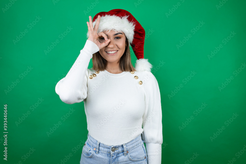 Young beautiful woman wearing a Santa hat and white sweater over green background doing ok gesture shocked with smiling face, eye looking through fingers Young beautiful woman wearing a Santa hat and white sweater over green background doing ok gesture shocked with smiling face, eye looking through fingers