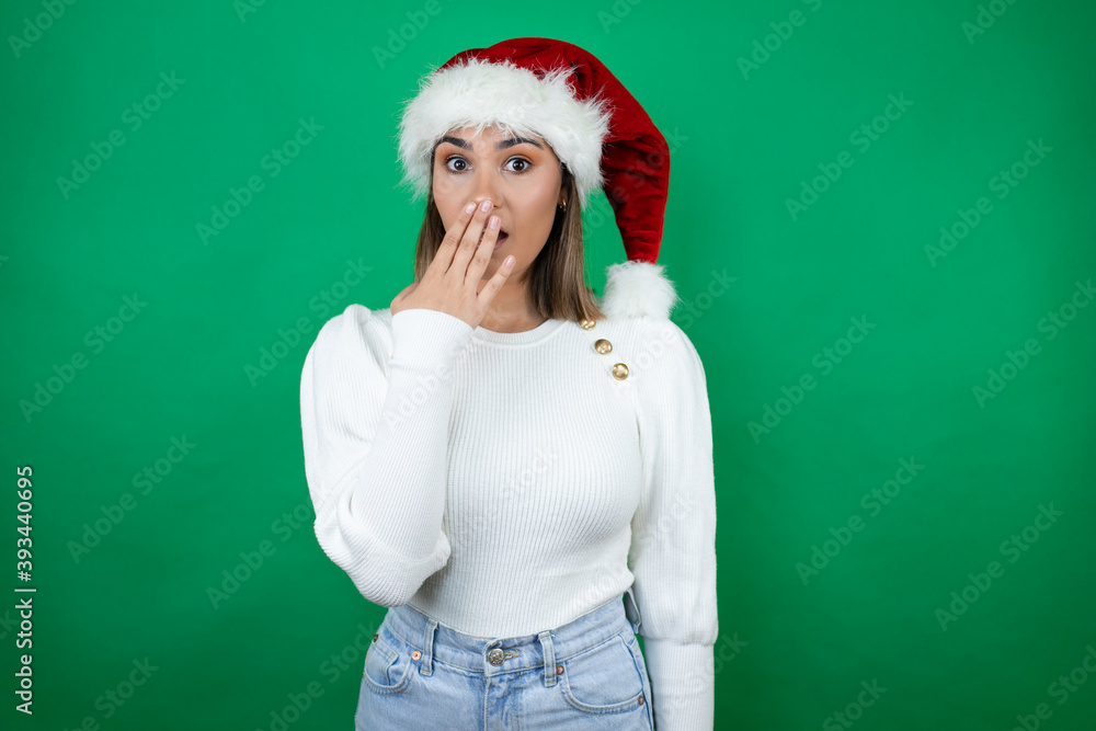 Young beautiful woman wearing a Santa hat and white sweater over green background surprised covering the mouth Young beautiful woman wearing a Santa hat and white sweater over green background surprised covering the mouth