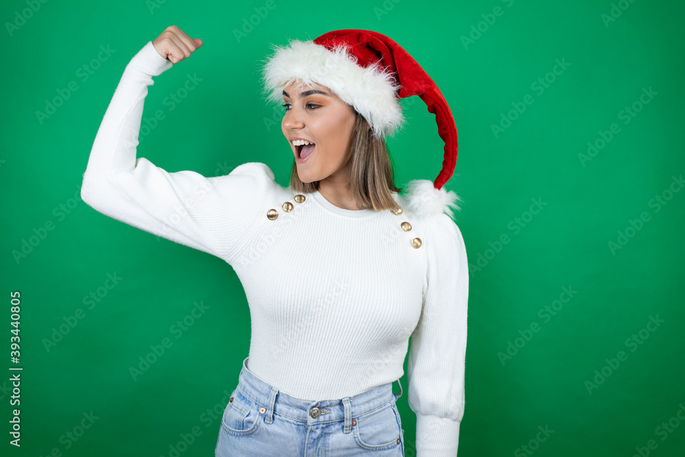 Young beautiful woman wearing a Santa hat and white sweater over green background showing arms muscles smiling proud Young beautiful woman wearing a Santa hat and white sweater over green background showing arms muscles smiling proud
