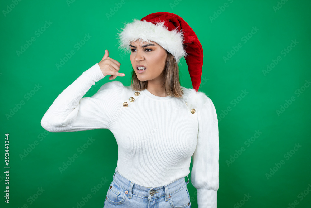 Young beautiful woman wearing a Santa hat and white sweater over green background confused doing phone gesture with hand and fingers like talking on the telephone Young beautiful woman wearing a Santa hat and white sweater over green background confused doing phone gesture with hand and fingers like talking on the telephone