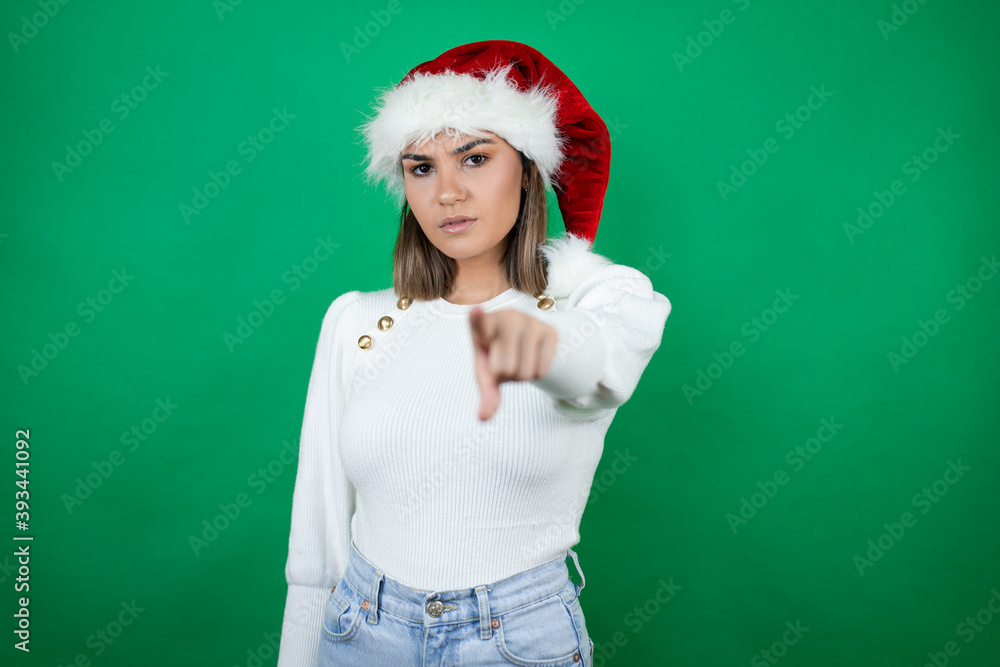 Young beautiful woman wearing a Santa hat and white sweater over green background pointing to the front with finger Young beautiful woman wearing a Santa hat and white sweater over green background pointing to the front with finger