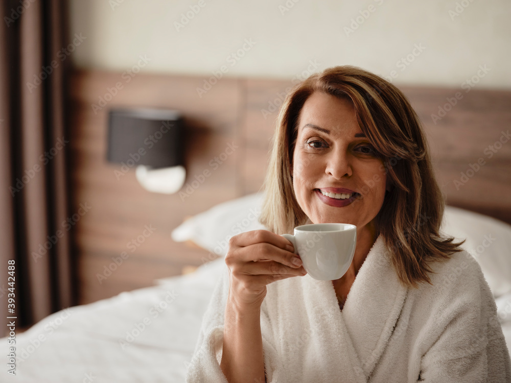 Smiling beautiful blond woman drinking coffee while sitting on bed at hotel room Smiling beautiful blond woman drinking coffee while sitting on bed at hotel room