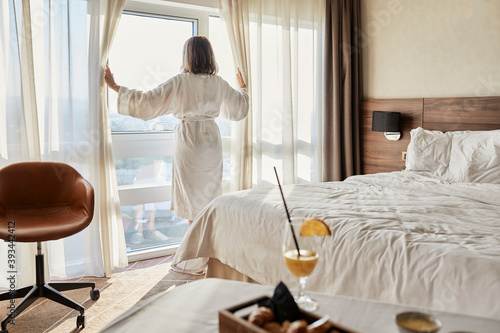Retired senior woman opening curtain while looking through window at luxury hotel room photo