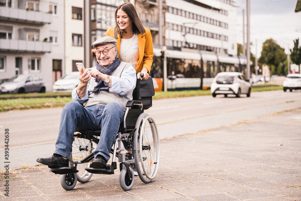 Smiling young woman pushing happy senior man with smartphone in wheelchair Smiling young woman pushing happy senior man with smartphone in wheelchair
