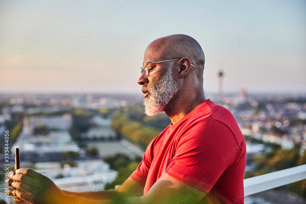 Bald man with white beard using smart phone for photographing sunset over city from terrace Bald man with white beard using smart phone for photographing sunset over city from terrace