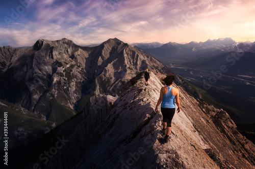Adventurous Girl is hiking up a rocky mountain. Dramatic Colorful Sunset Sky. Taken from Mt Lady MacDonald, Canmore, Alberta, Canada. Epic Adventure Travel
